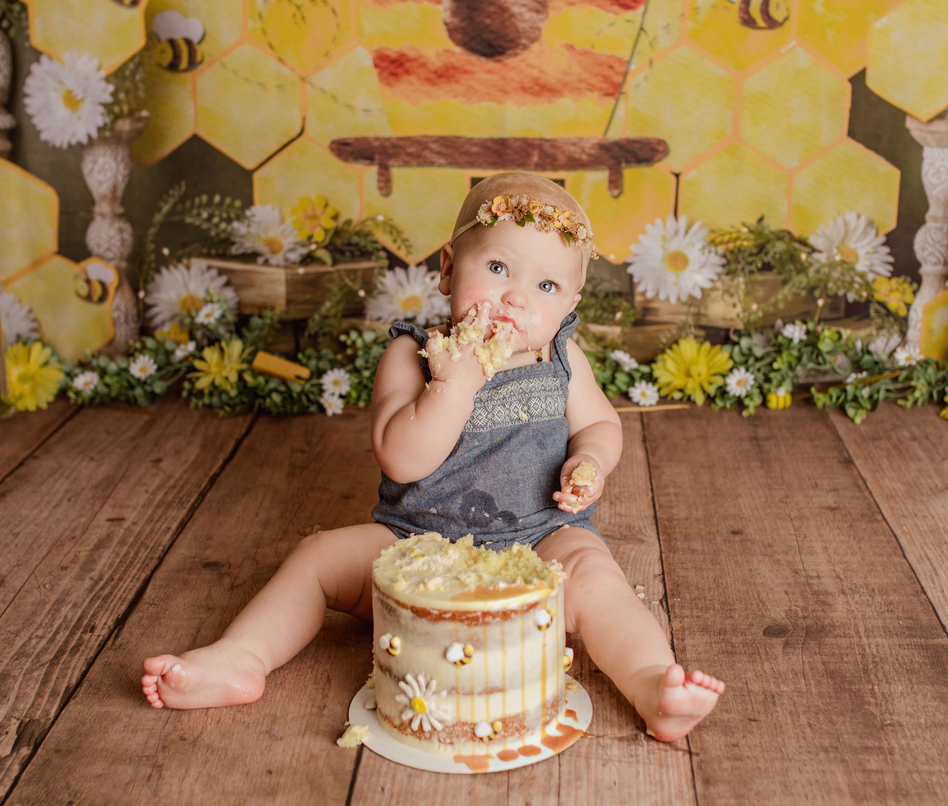Toddler eating cake during a bee-themed first birthday photo shoot; yellow honeycomb backdrop, wooden floor.