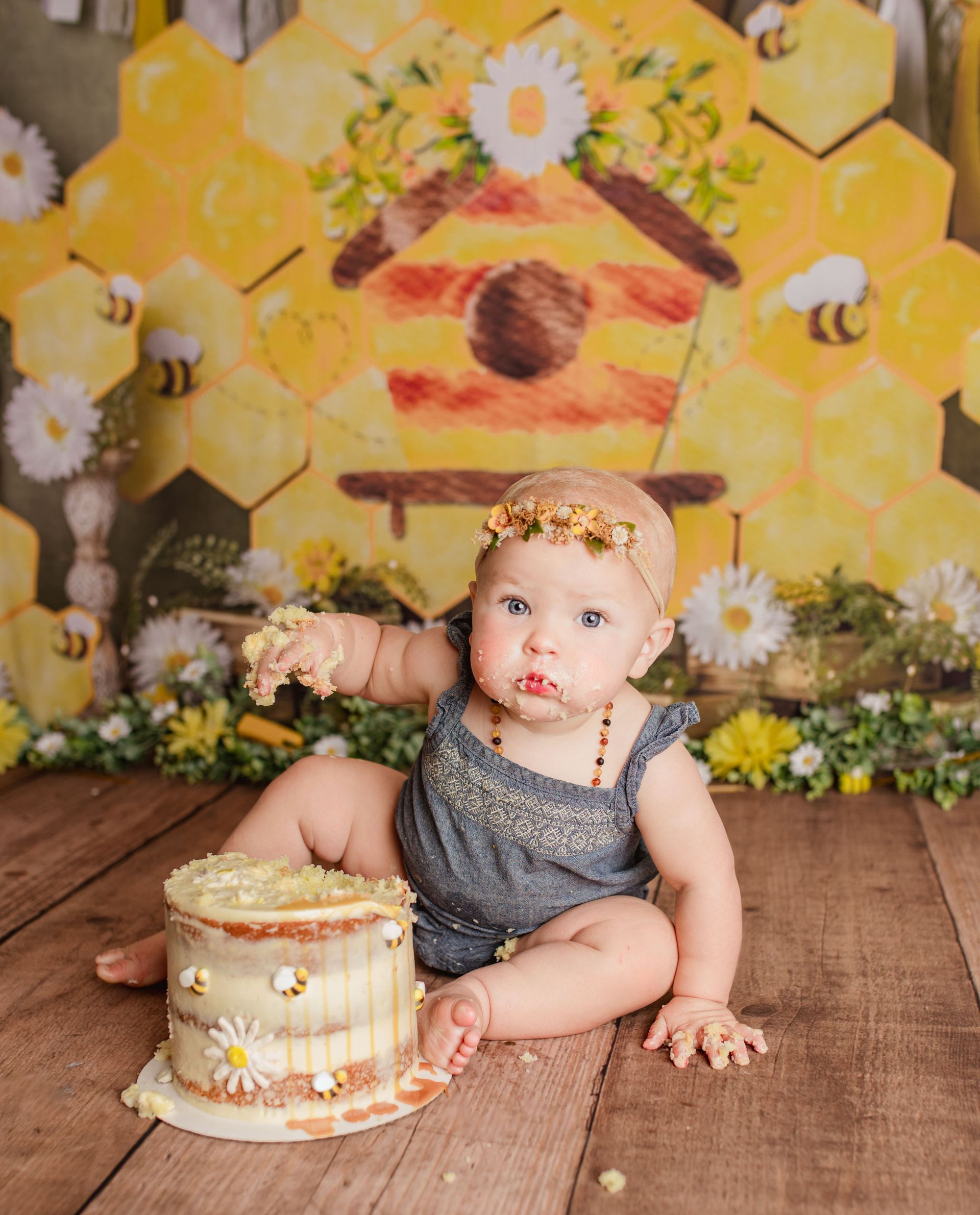 Baby with messy face sits by a cake on wooden floor, bee-themed backdrop.
