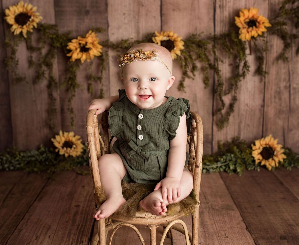 Smiling baby in a green outfit sits in a wicker chair, surrounded by sunflowers against a wood background.