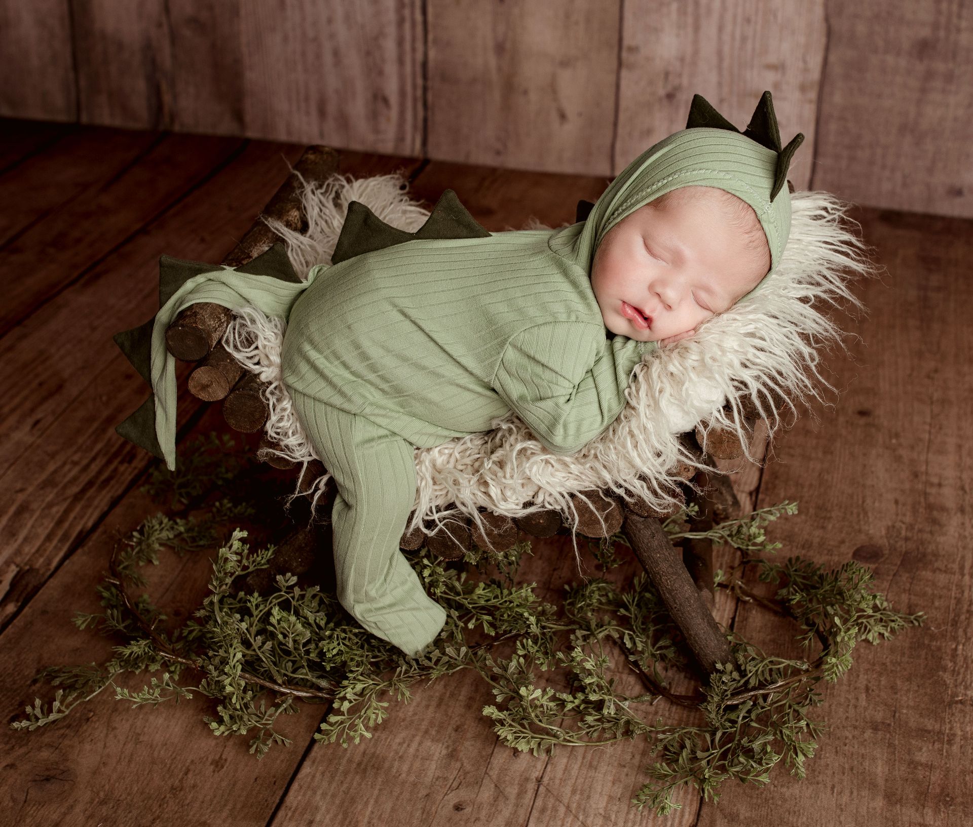 Newborn baby asleep in a dinosaur costume, resting in a wooden basket with fur and greenery.