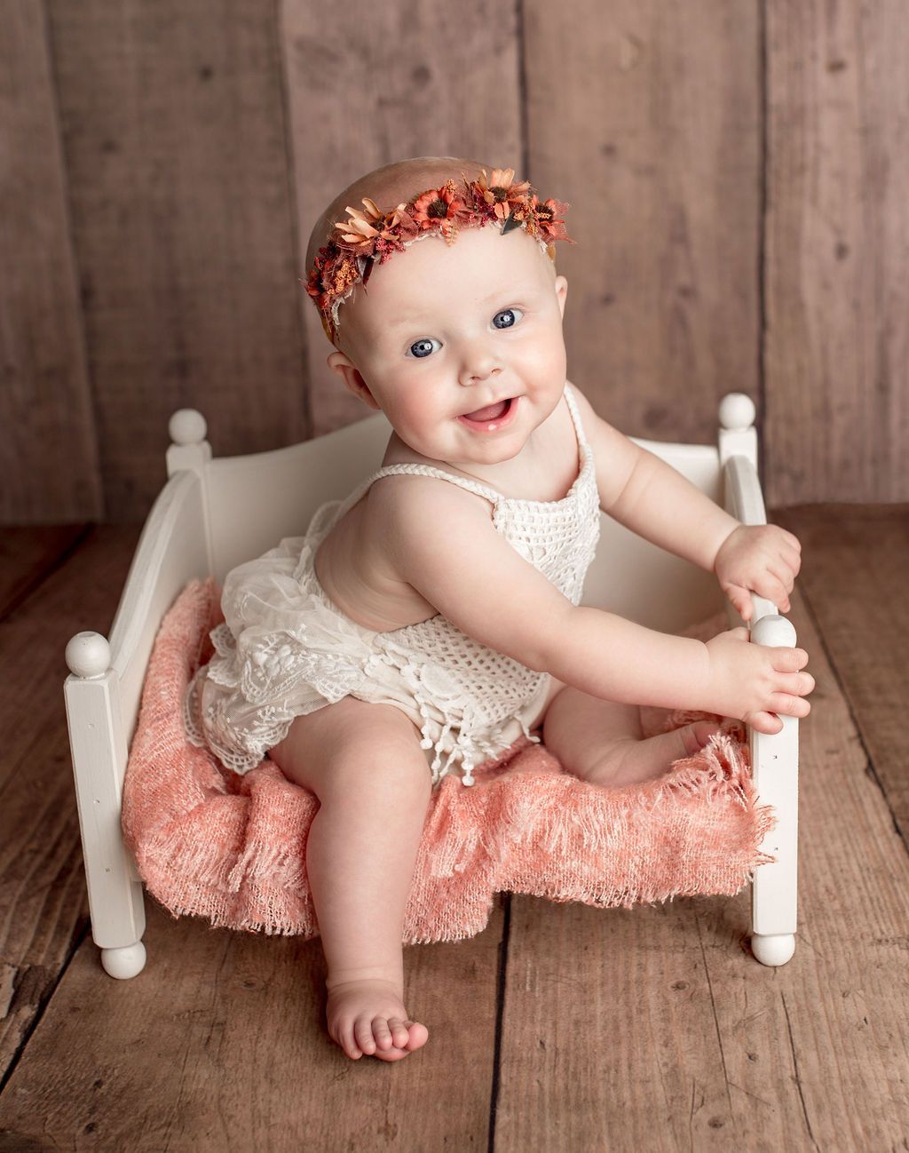 Smiling baby in white lace outfit and flower crown sits in small white bed.