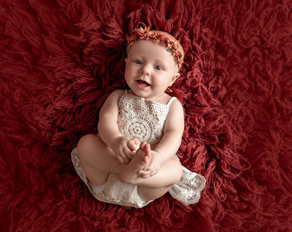 Baby in crochet outfit, floral headband, smiling on red fur blanket.