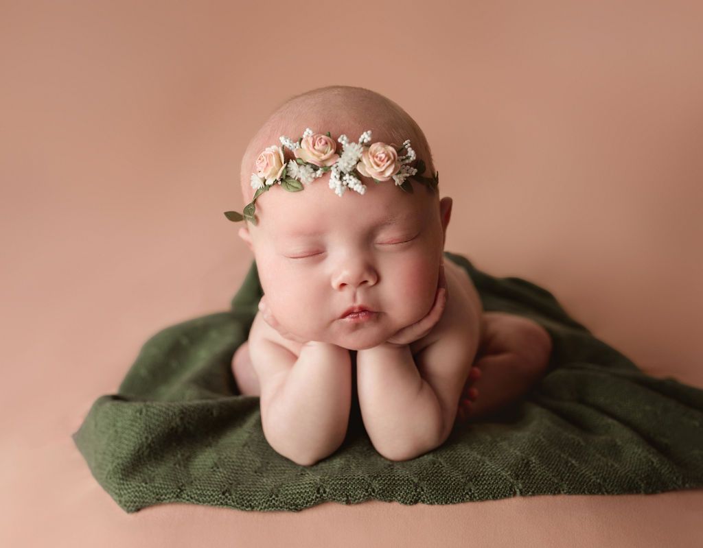 Baby with floral crown rests on hands, eyes closed, on a green blanket against a peach backdrop.