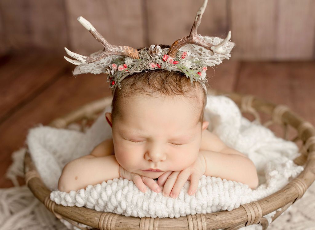 Newborn in basket, wearing antler headband, resting on white blanket.