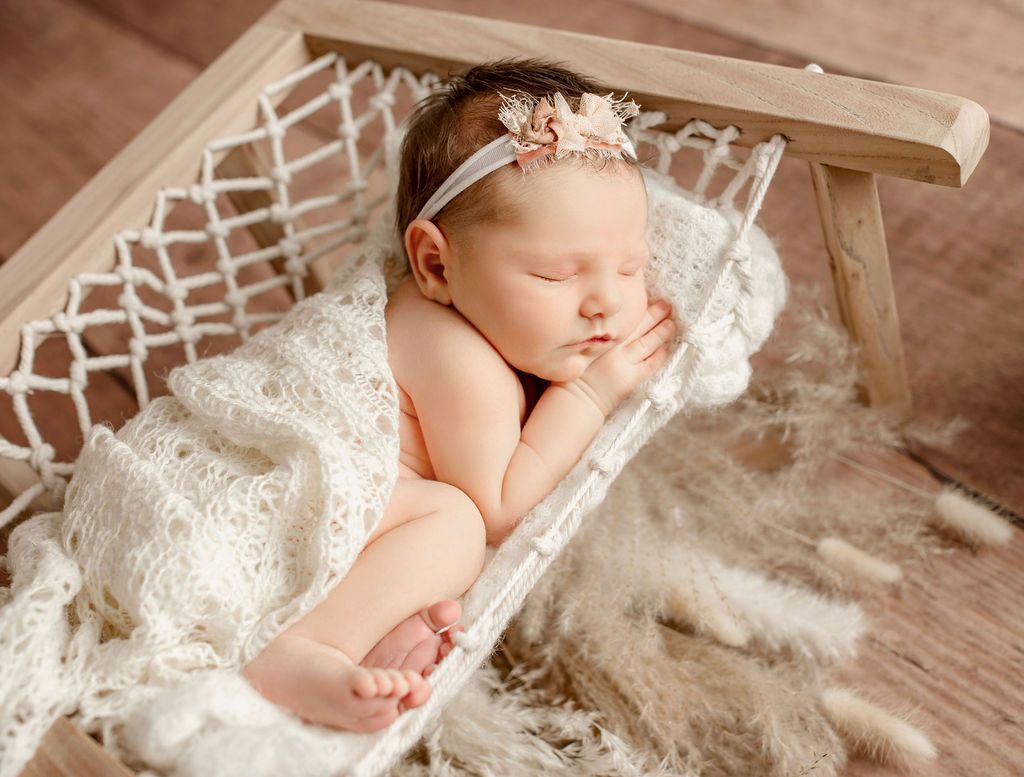 Newborn asleep in a small wooden hammock, wrapped in a white blanket. White headband with a flower.