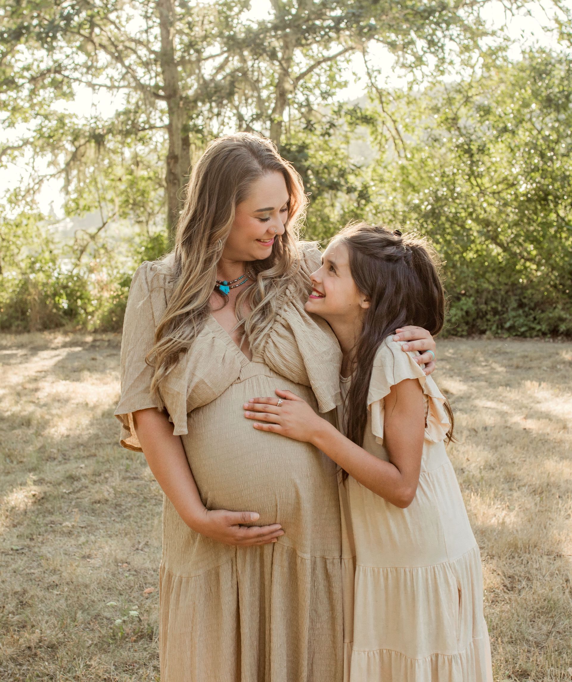 Pregnant woman and girl embrace outdoors, looking at each other. Both wearing light dresses.