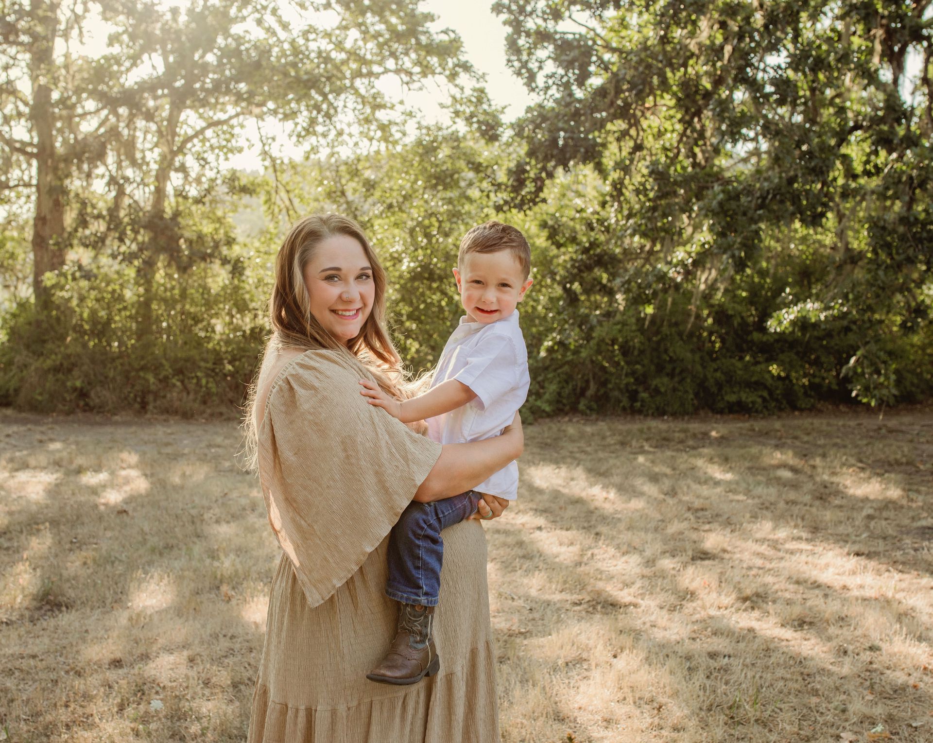 Woman holding a child outdoors, smiling. Both are in front of trees in sunlight.