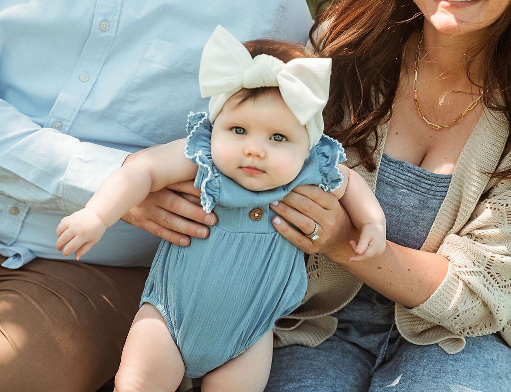 Baby in blue romper and cream bow headband, held by parents.