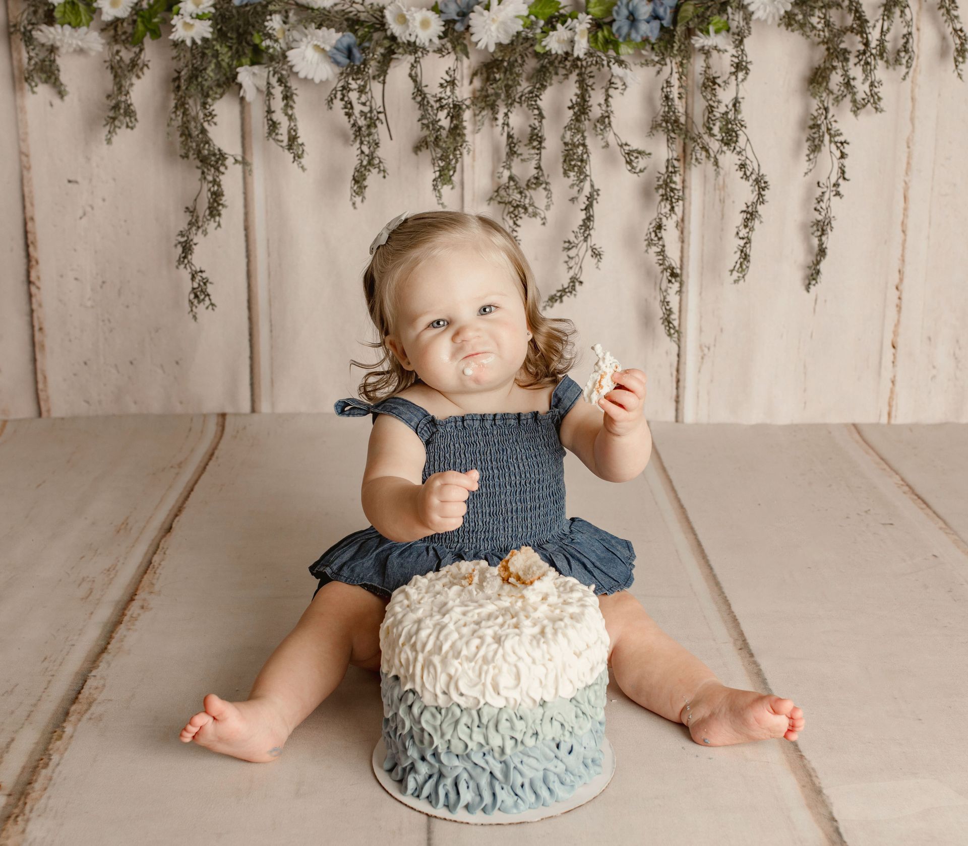 A baby in a blue dress sits with a frosted cake, messy-faced and holding a piece. Wooden backdrop.