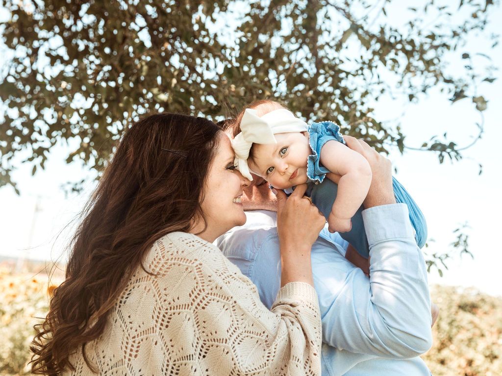 Parents holding a baby. The baby, in a denim outfit, is lifted up. They're all smiling outdoors.