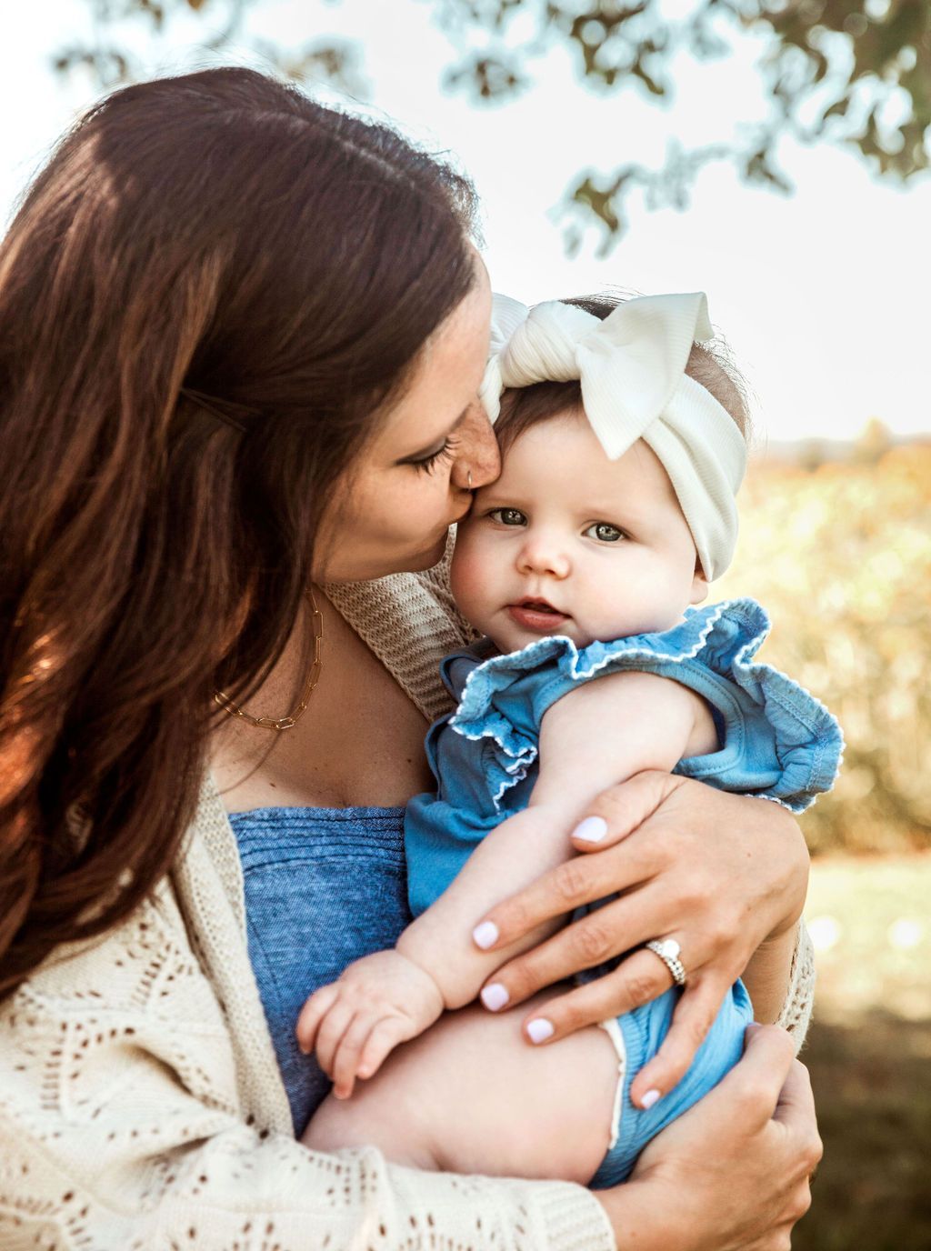 Woman kisses baby's forehead. Both are outdoors. Baby wears blue romper and bow headband.
