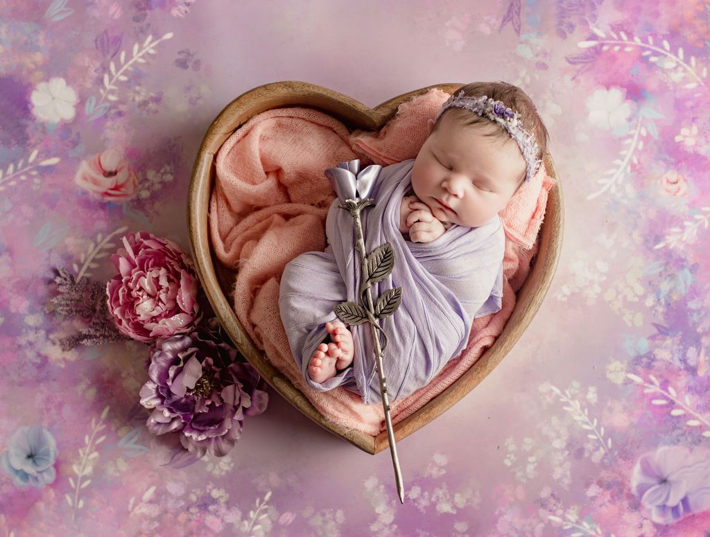 Newborn baby swaddled in purple blanket, resting in a heart-shaped wooden bowl, holding silver rose.