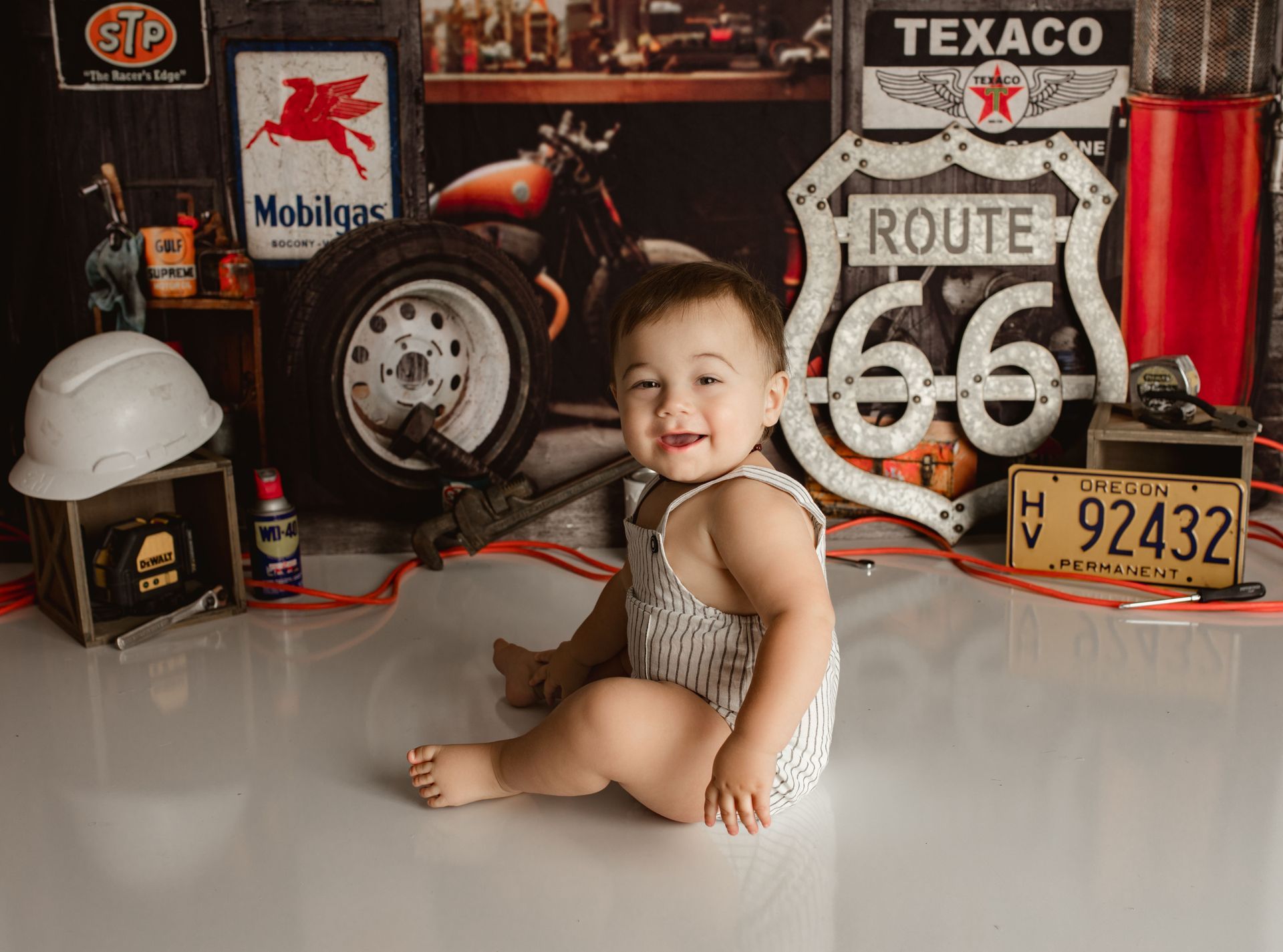 Smiling baby in striped onesie, sitting on floor in front of Route 66 and garage-themed backdrop.