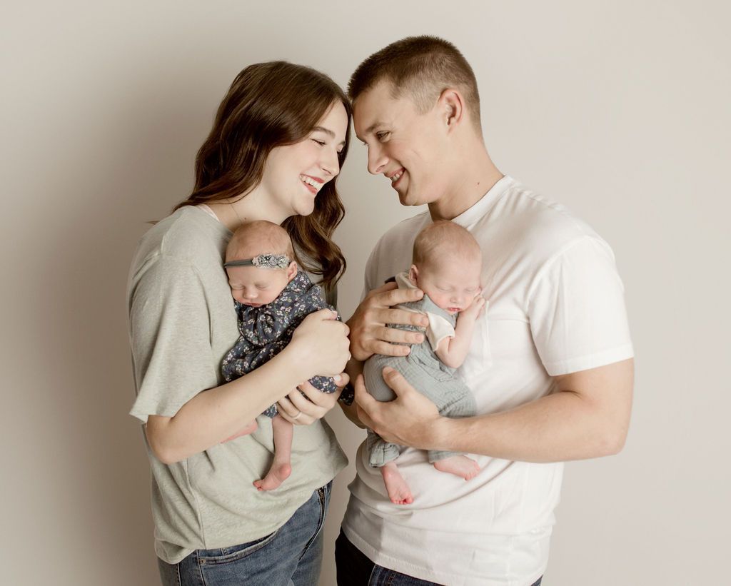 Couple smiles while holding newborn twins against a light background.
