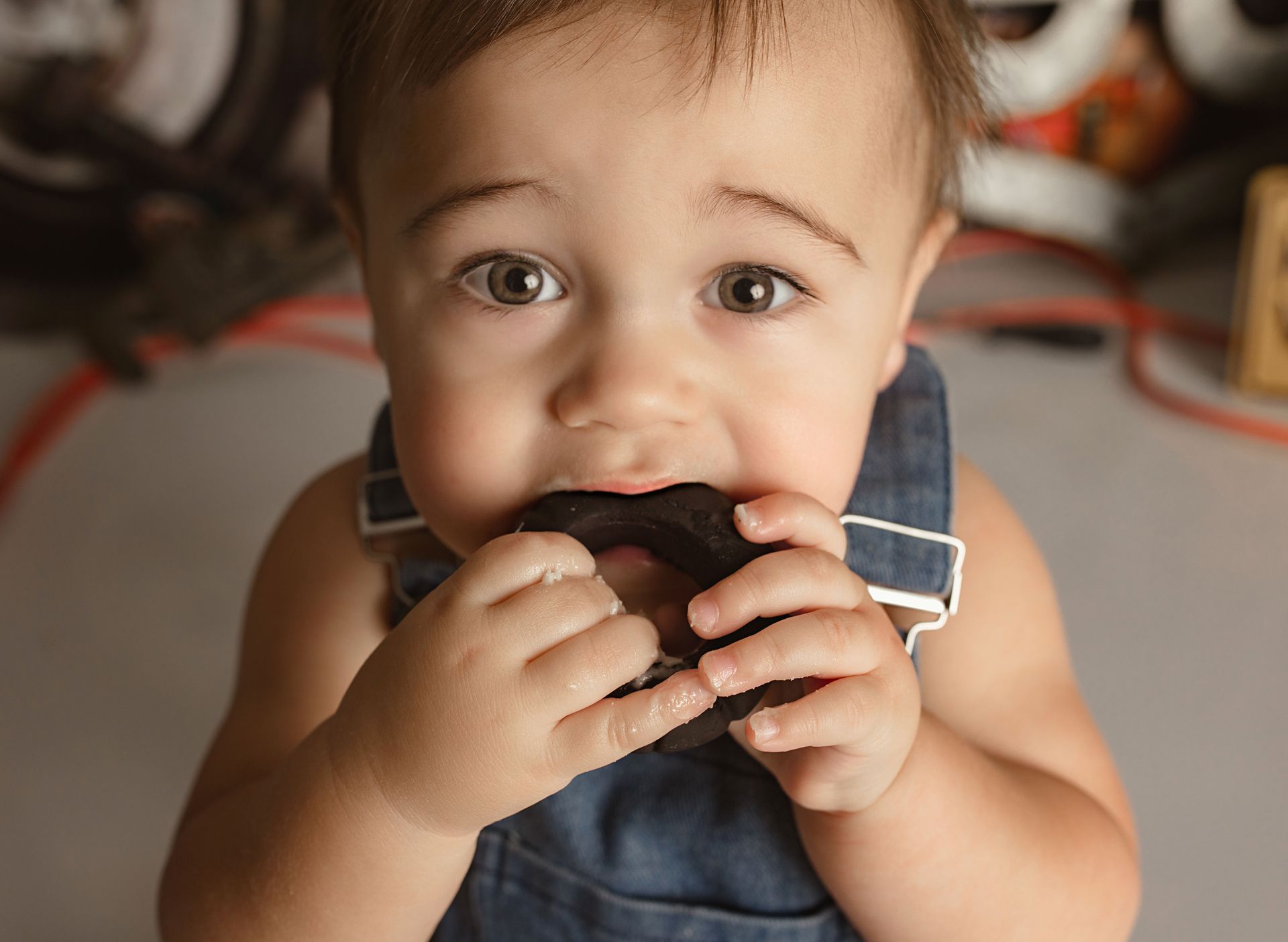 Baby chewing on a black object, wearing blue overalls, looking up with wide eyes.