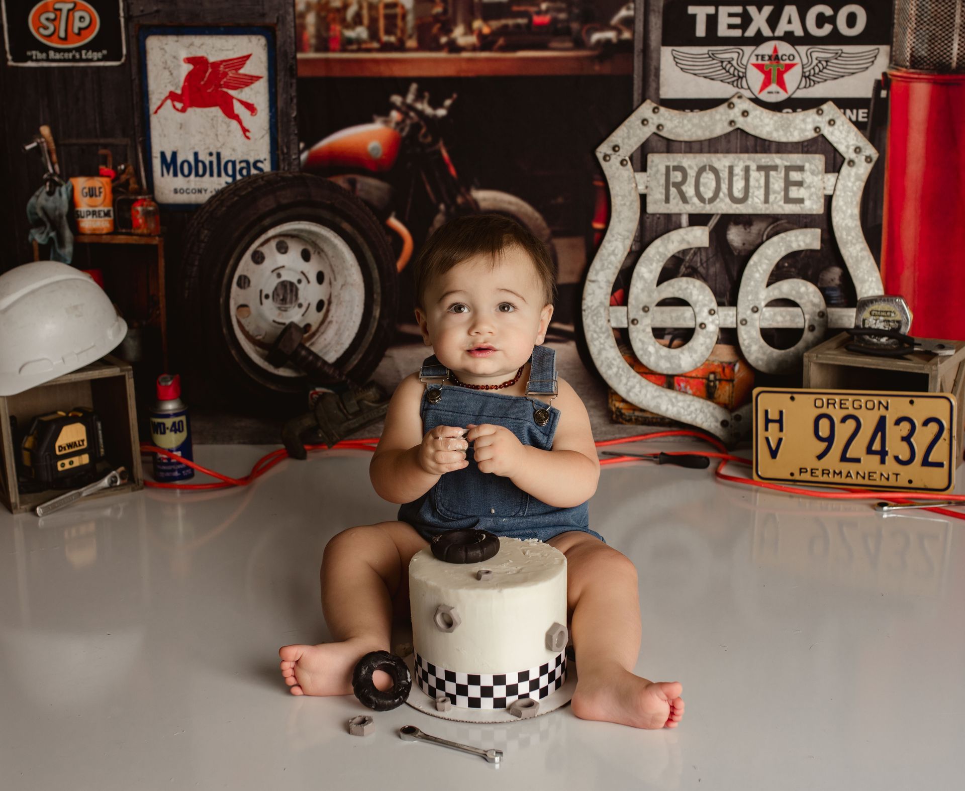 A baby sits on a checkered cylinder surrounded by tools and auto-themed decor.