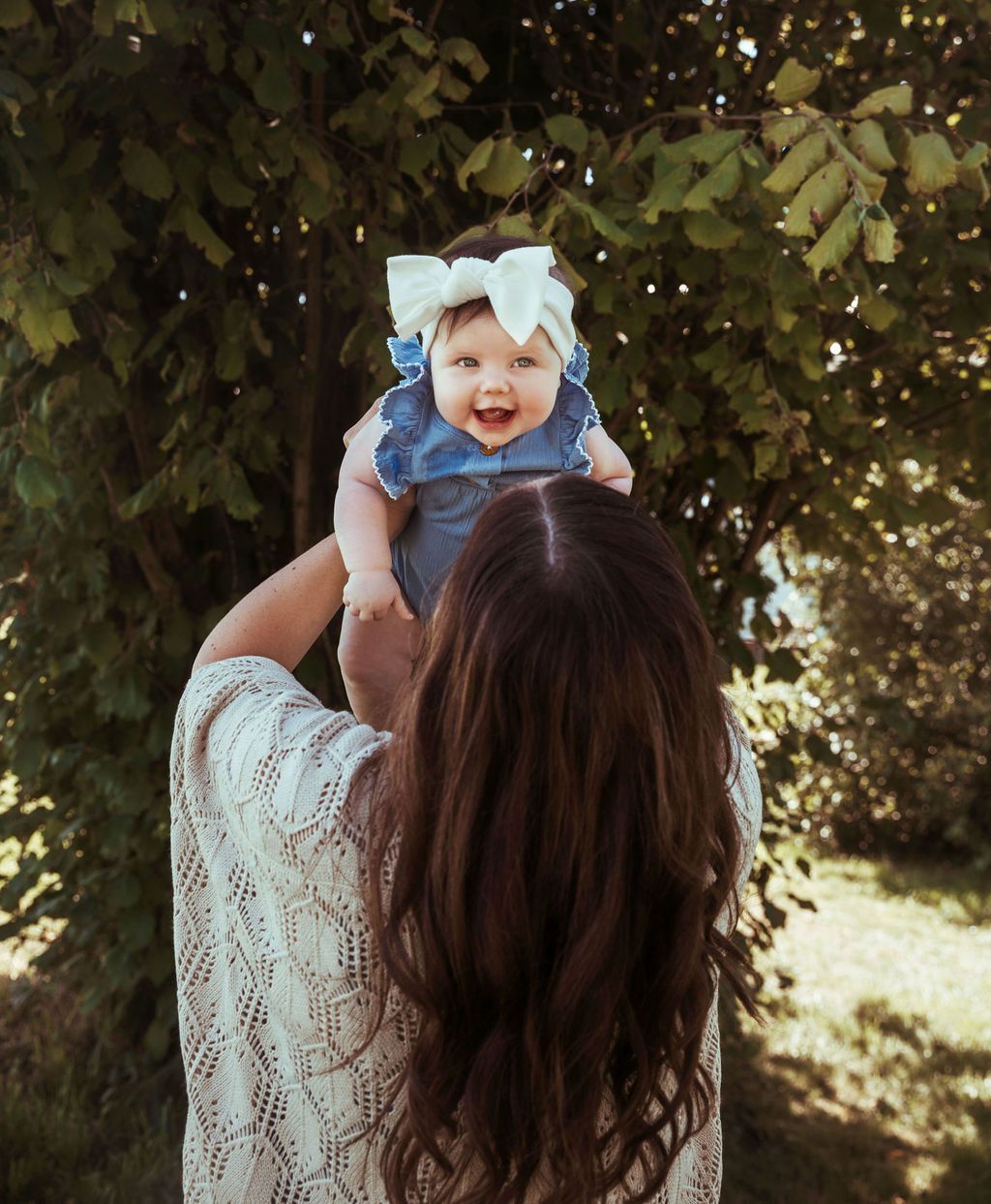 Woman holding a smiling baby up in the air. Both are outdoors near a tree. Baby wears a bow.