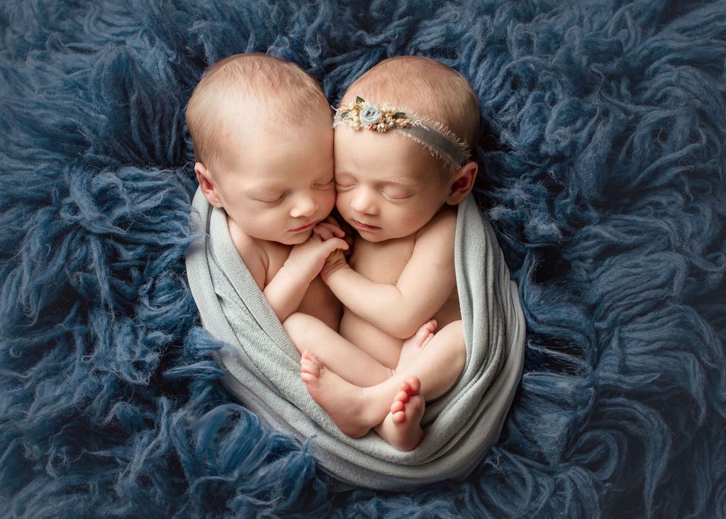 Newborn twins nestled in a blue wrap, lying on blue fluffy fabric, heads touching, one with floral headband.