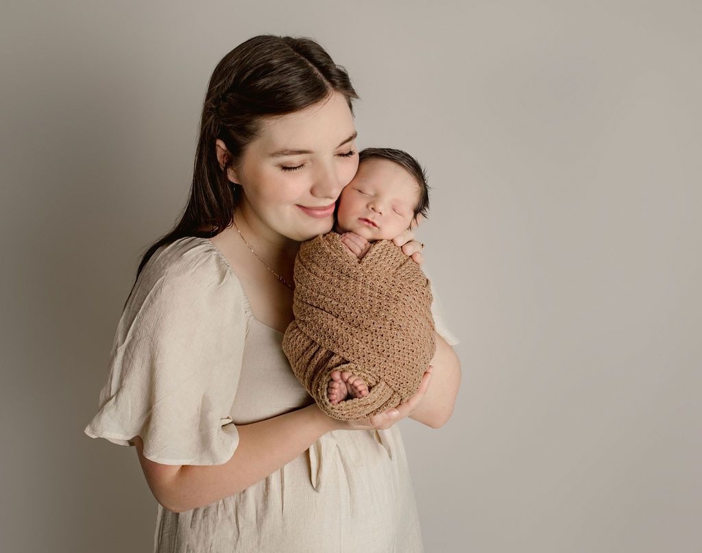 Woman holding a swaddled newborn, both with eyes closed, against a gray background.