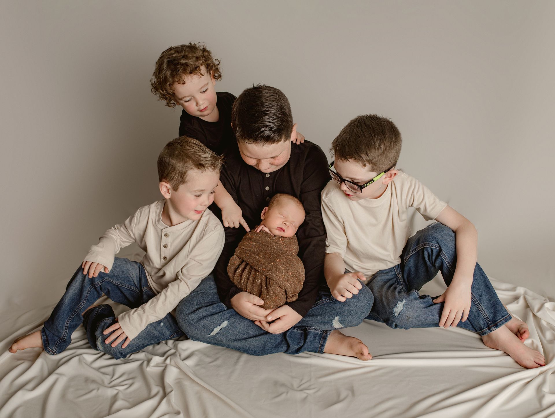 Five children surround a newborn, looking down. They sit on a white cloth in a studio setting.
