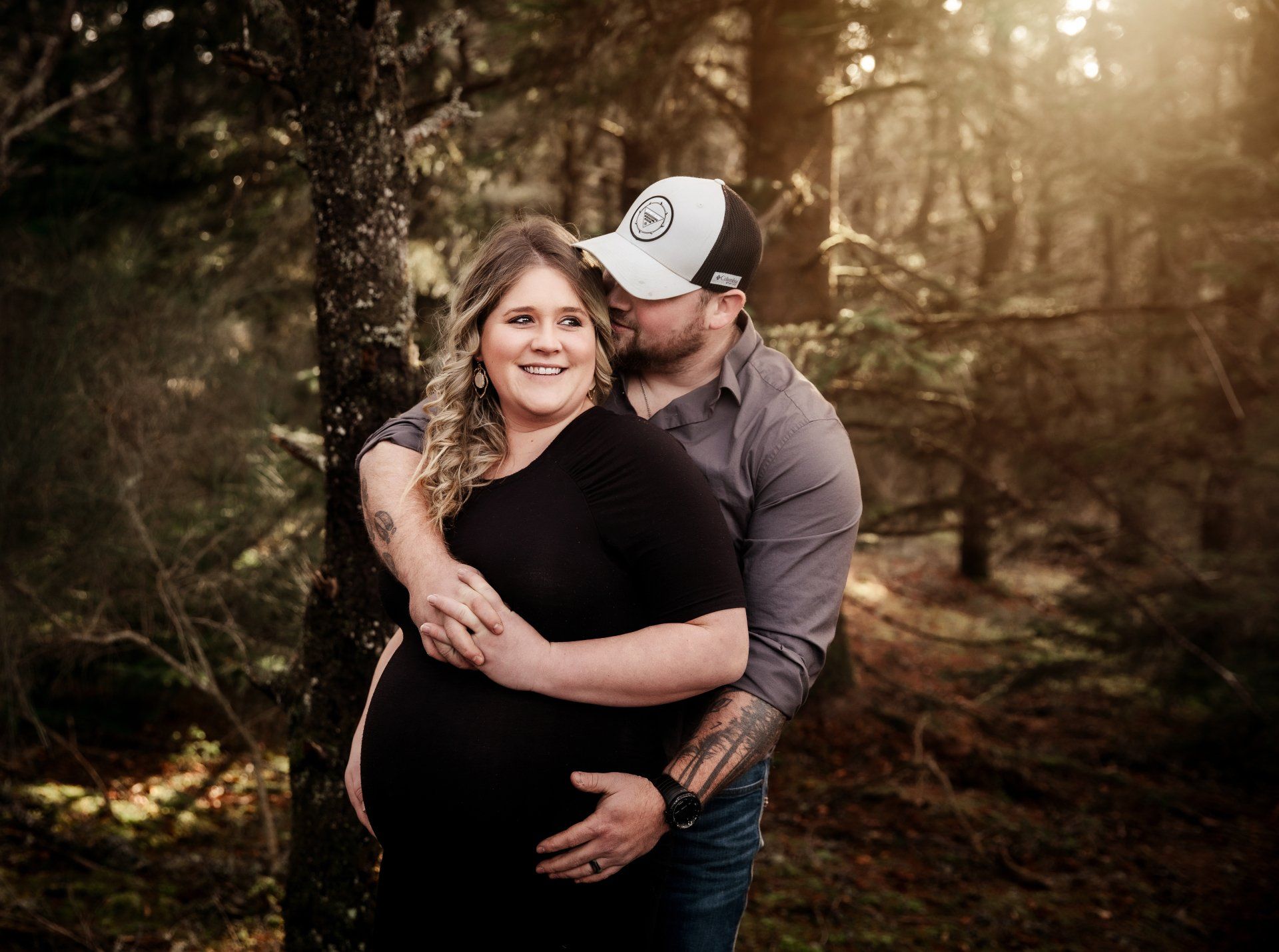 Pregnant woman in a black dress, embraced by a man in a forest, both smiling.