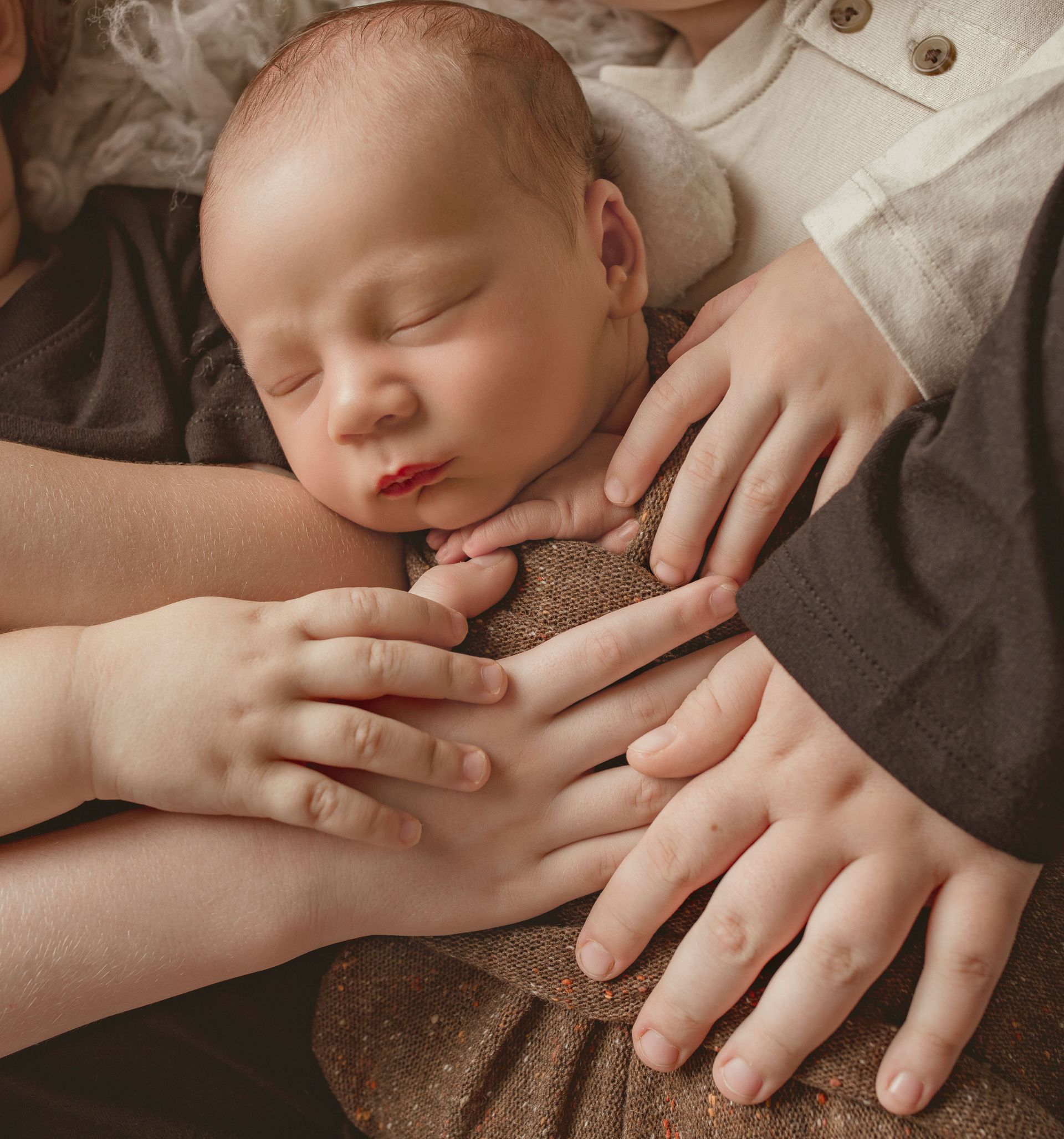 Sleeping baby held gently by multiple hands, wearing a brown onesie.