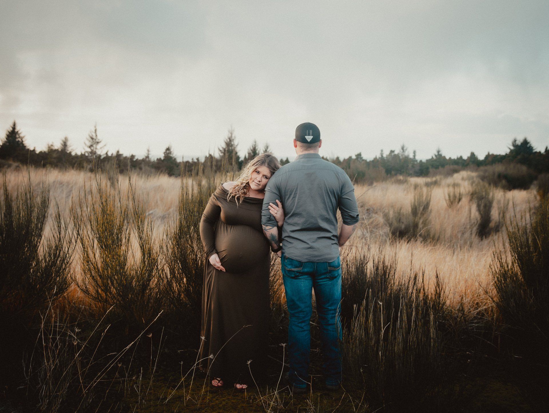 Pregnant person in brown dress leans on a person wearing a hat and blue jeans, standing in tall grass.