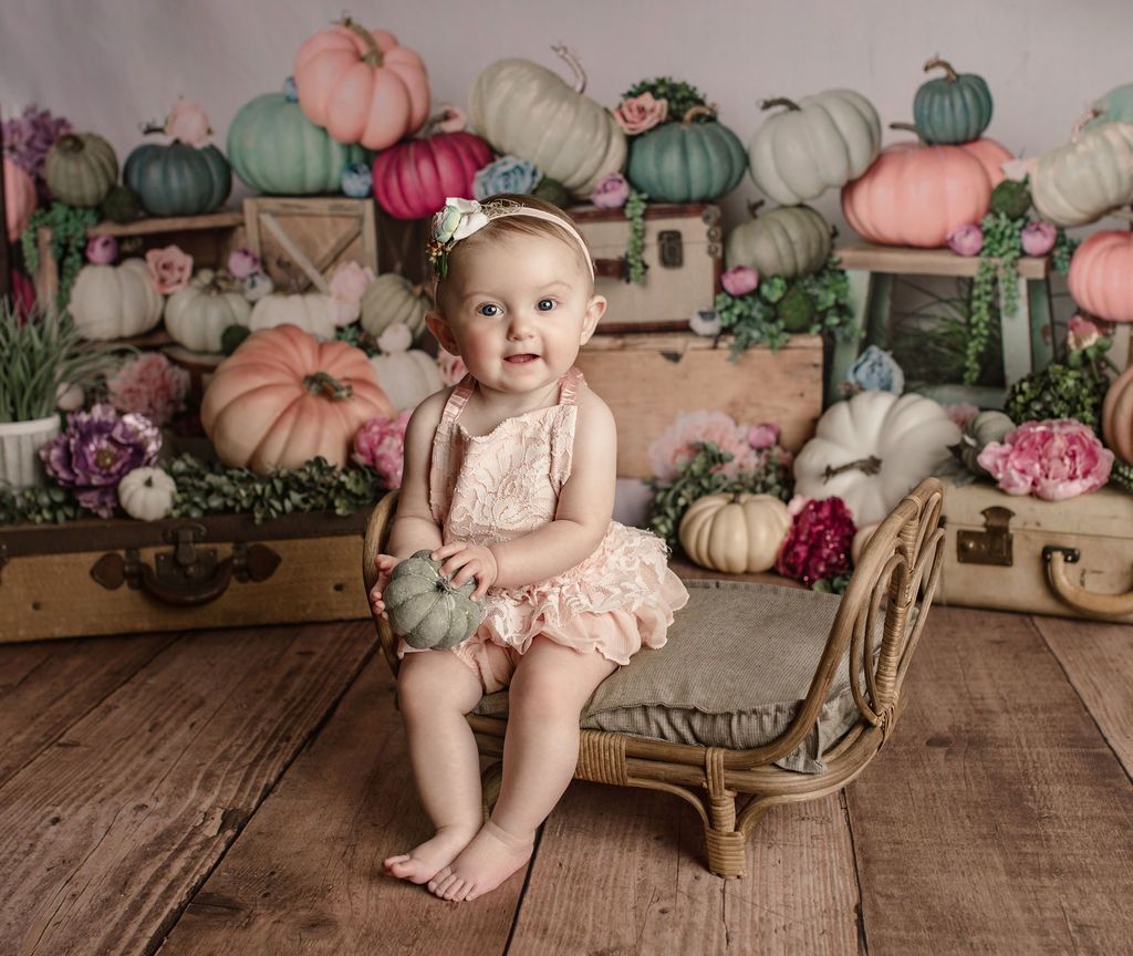 Baby sitting on a small bench, holding a small pumpkin, surrounded by pumpkins and flowers, wooden backdrop.