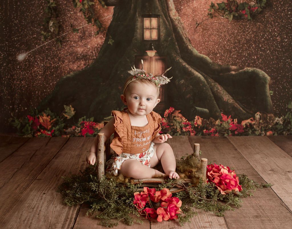 Baby in a whimsical forest-themed setting, seated on a miniature rustic bench, looking at the viewer.