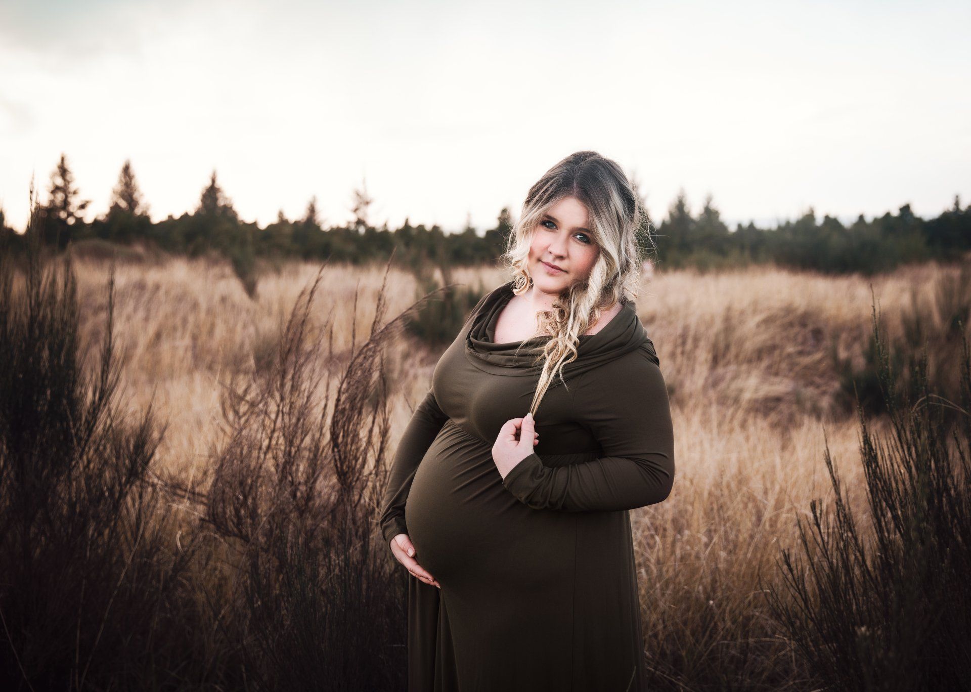 Pregnant person in olive dress poses in a field, holding belly, golden hour.
