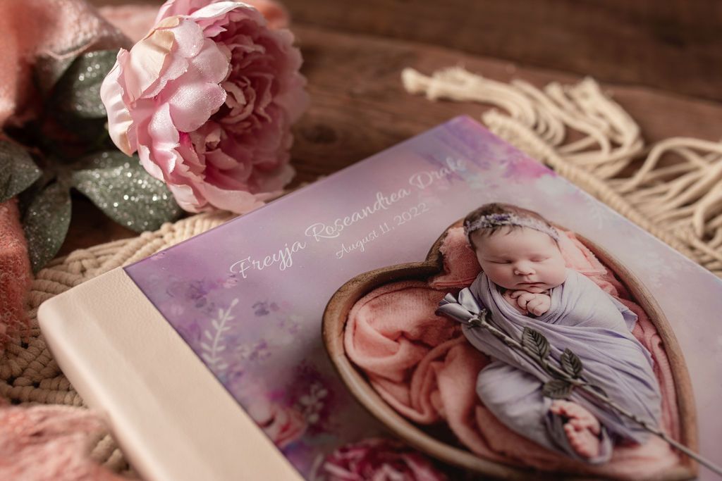 A newborn in a heart-shaped prop, on the cover of a photo album with a pink peony.
