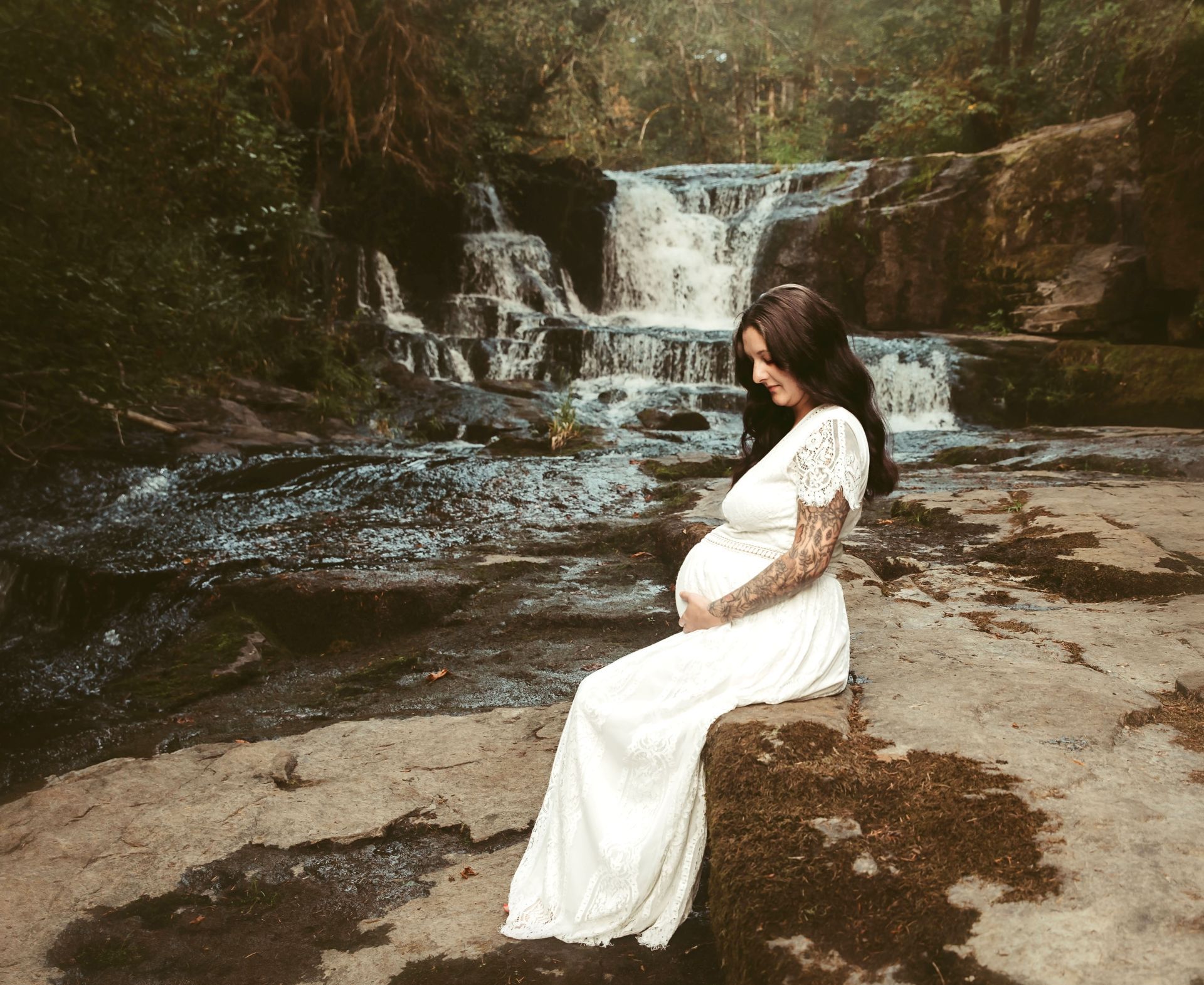 Pregnant person in white dress sits on a rock, holding belly, with waterfall backdrop.