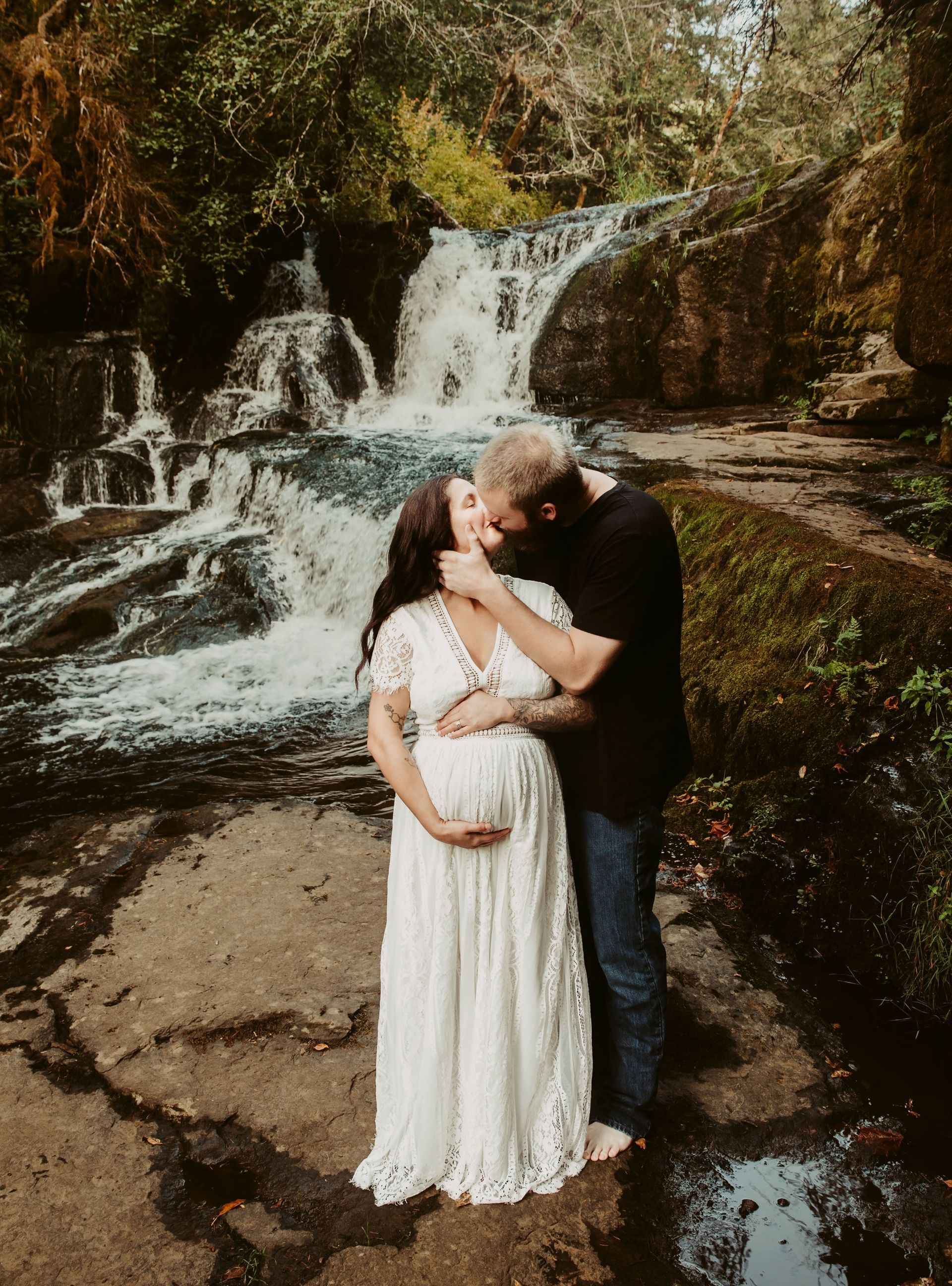 Pregnant couple kissing in front of a waterfall; woman in white dress, man in black shirt.