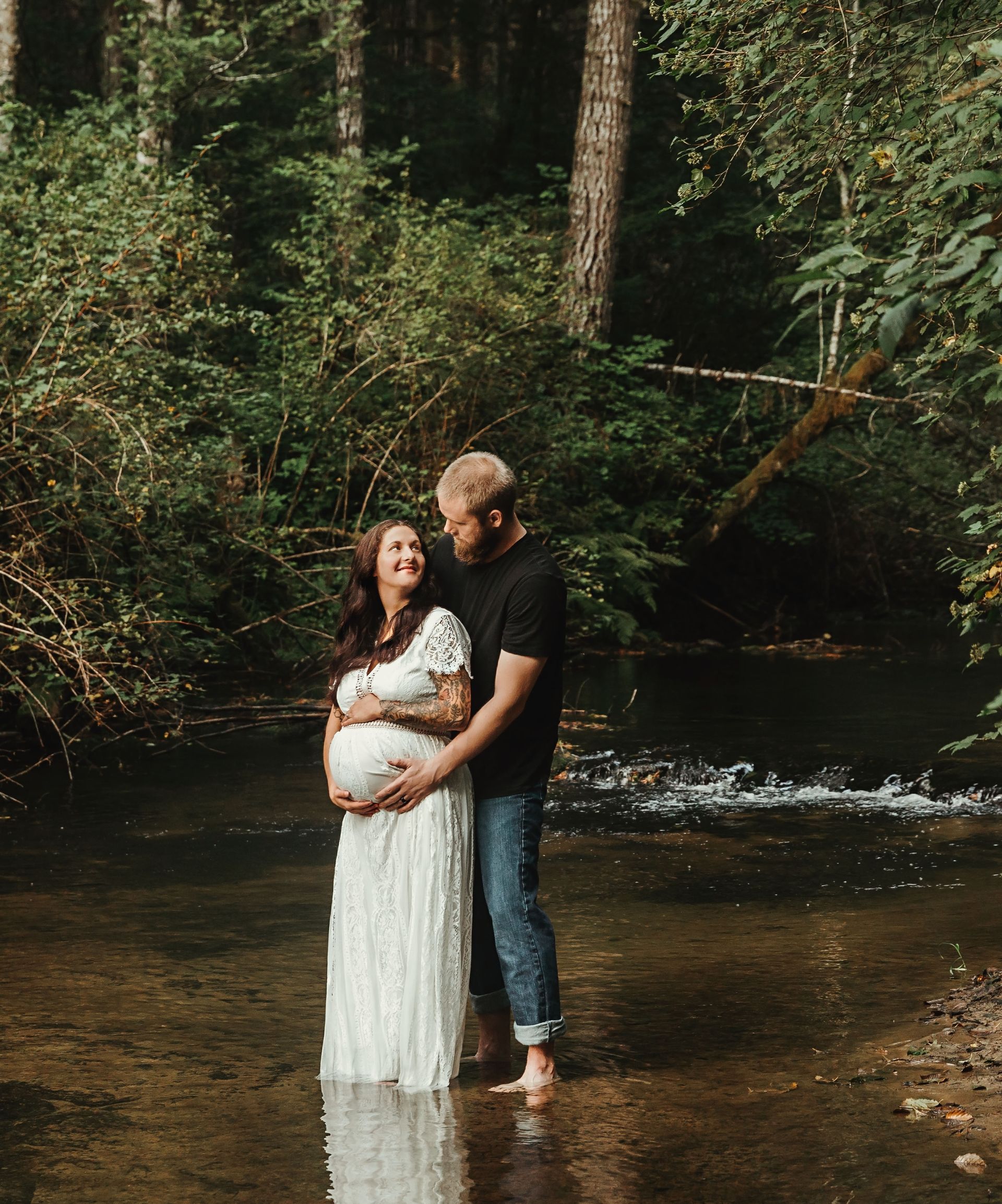 Couple embracing in a shallow stream, woman's pregnant belly visible. Forest setting.