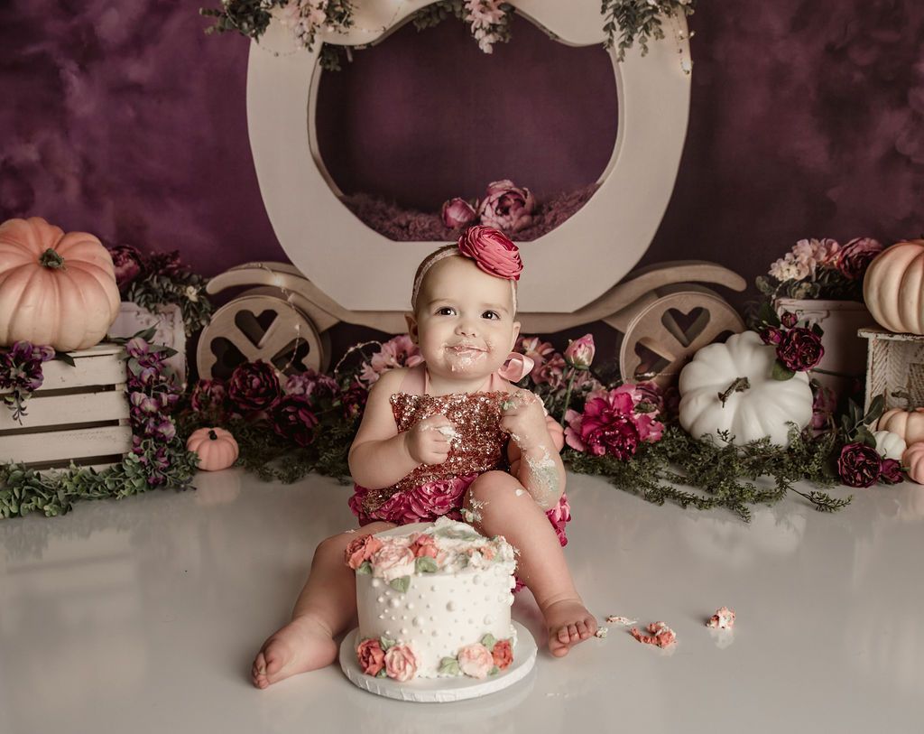 Baby in pink floral outfit sits with cake; smeared frosting on face and body. Pumpkin and floral background.