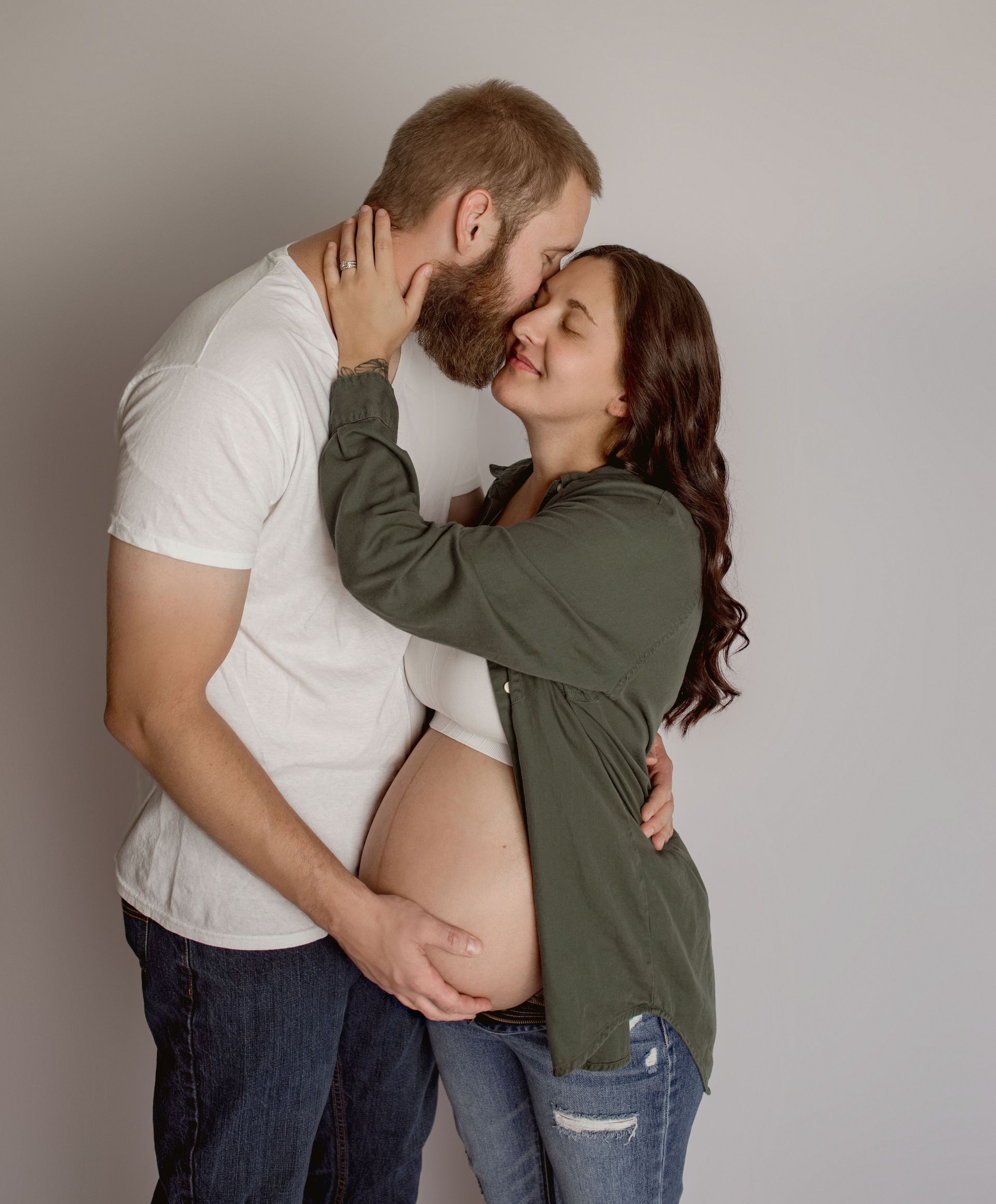Pregnant woman embraces man, both smiling. He kisses her forehead, hand on her belly.