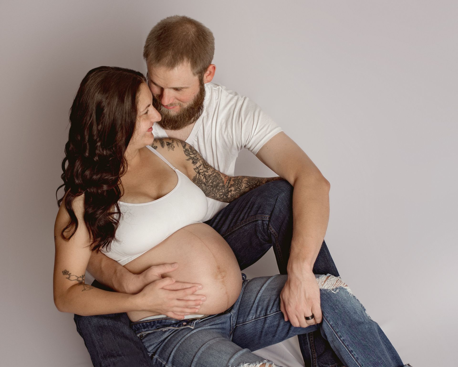 Pregnant woman and partner, sitting close, man's arms around her, white top, denim, studio.