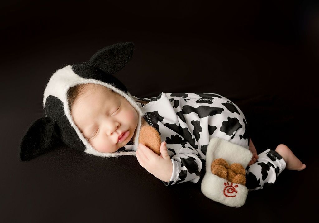 Baby in cow costume sleeping, holding a Chick-fil-A chicken nuggets toy. On a black background.