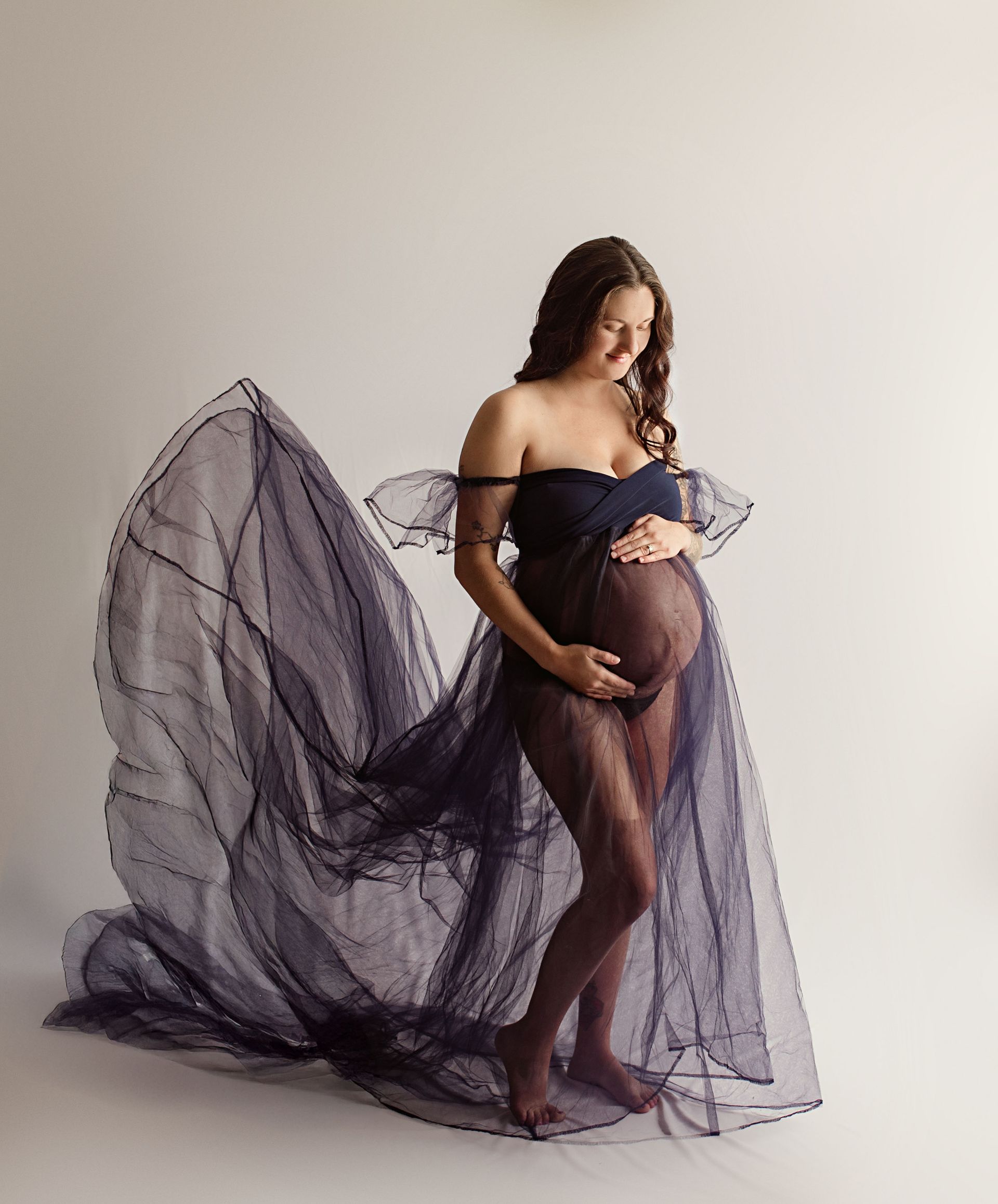 Pregnant person in dark blue sheer gown, holding belly, against a light backdrop.