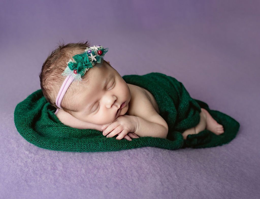 Newborn baby asleep, wrapped in a green blanket, wearing a floral headband on a purple surface.