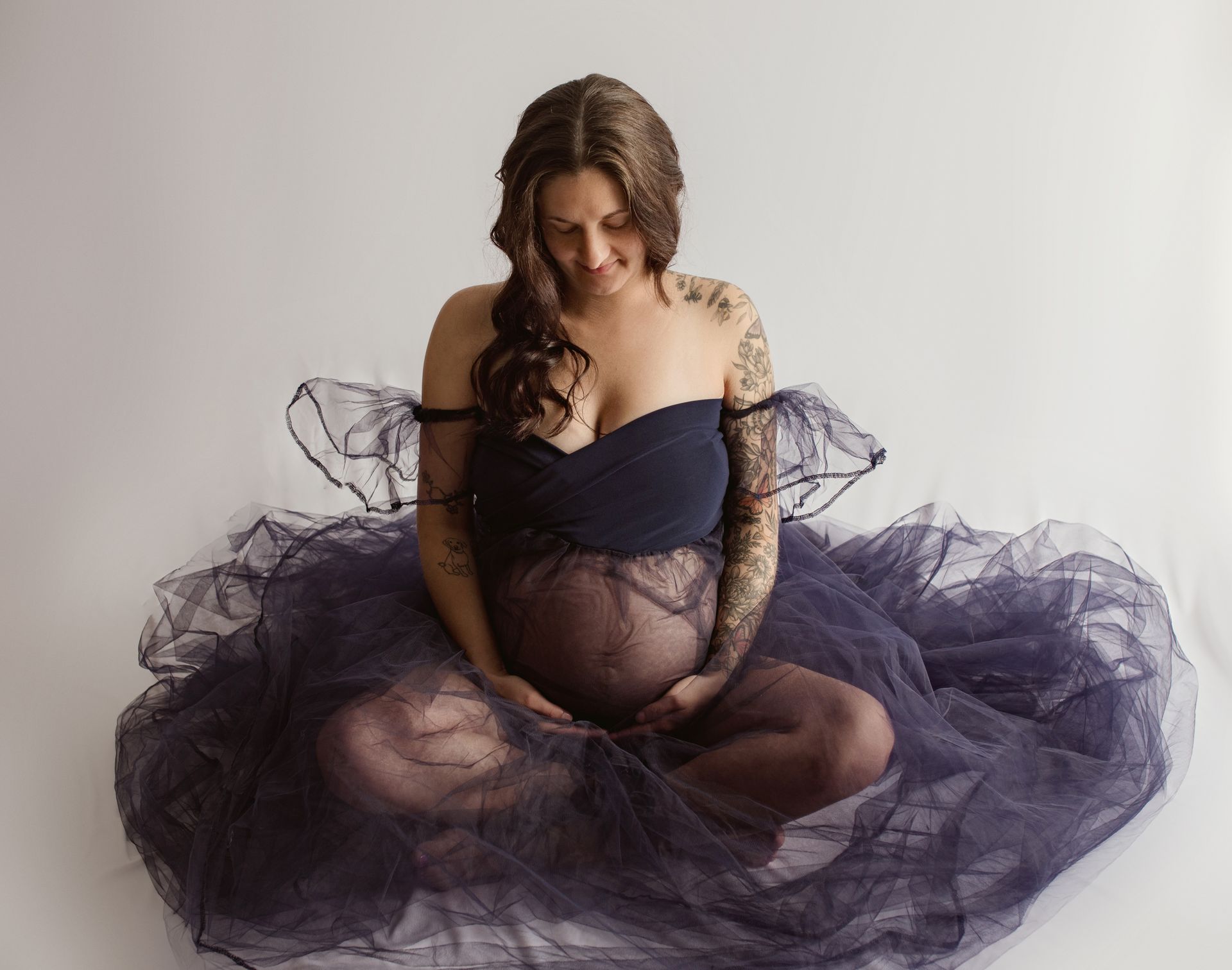 Pregnant person in navy tulle dress sits cross-legged, holding belly, looking down.