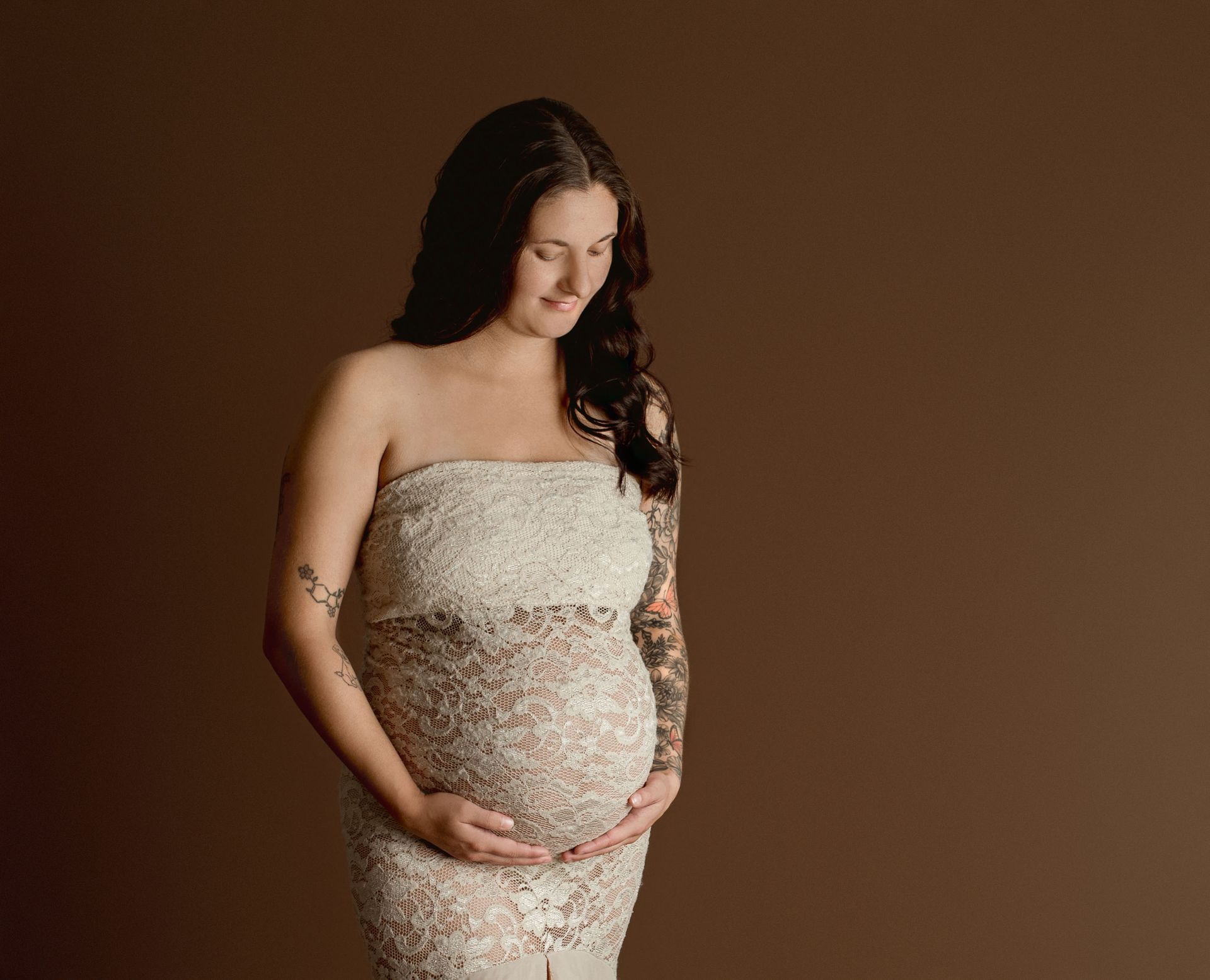 Pregnant woman in a white lace dress holds her belly. Brown background.