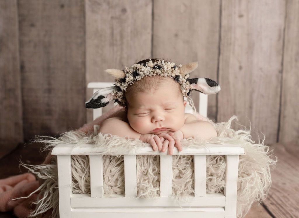 Newborn baby wearing cow hat asleep on small white bed.