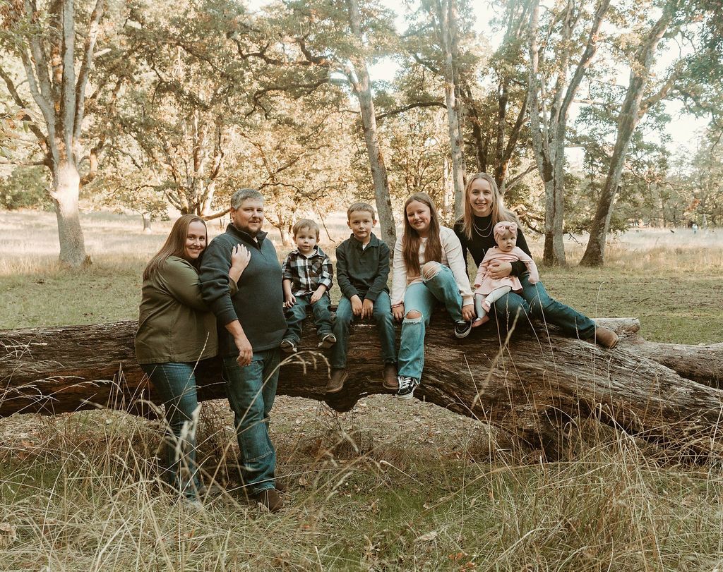 Family of eight sitting on a large fallen log in a park-like setting.