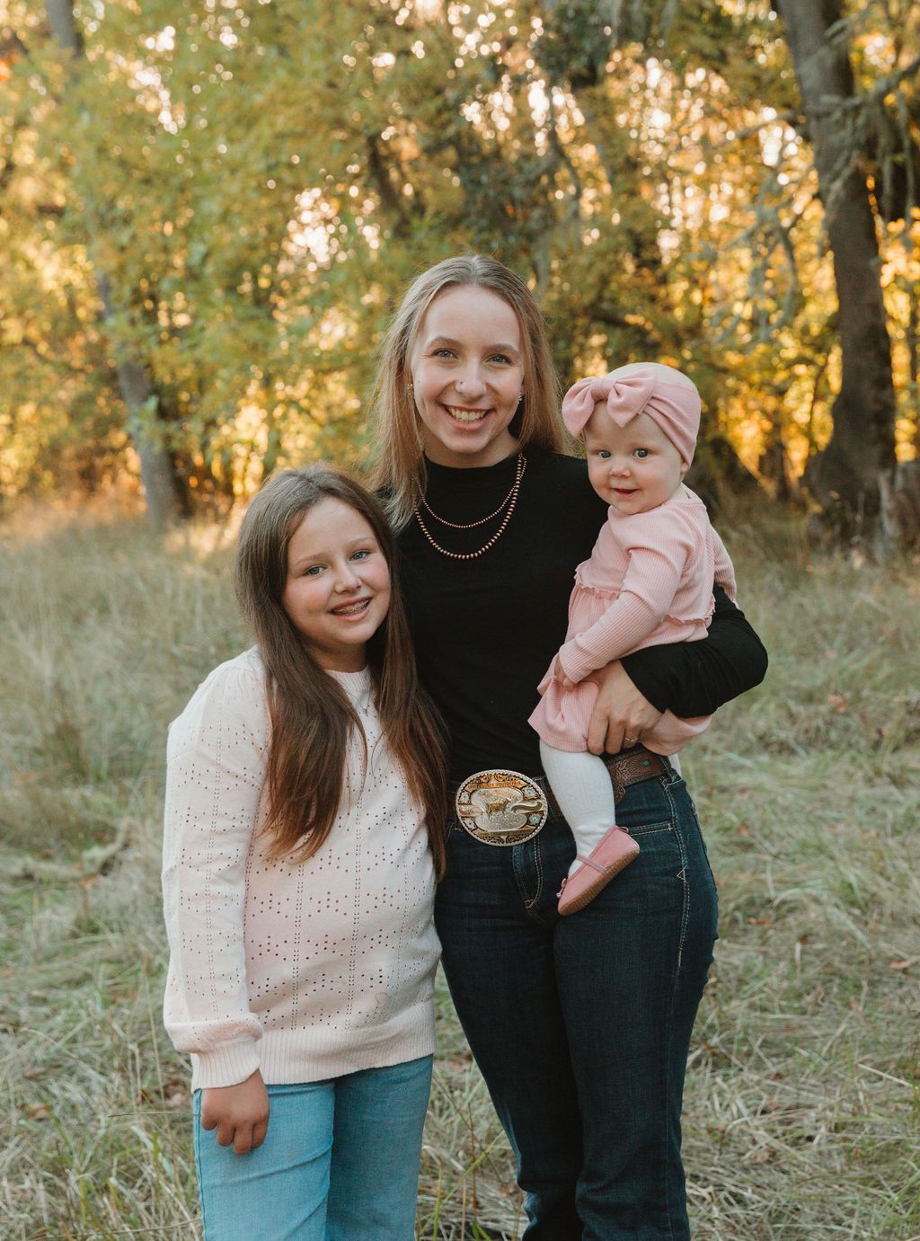 Woman holding a baby, standing with a girl in a field with trees. All are smiling.