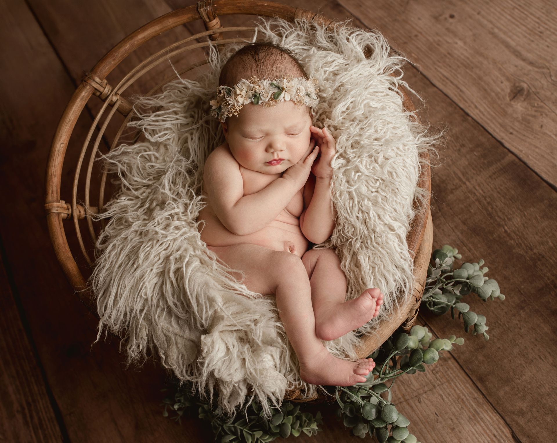Newborn sleeping in a woven basket with fluffy blanket, wearing a floral crown, on wooden surface.