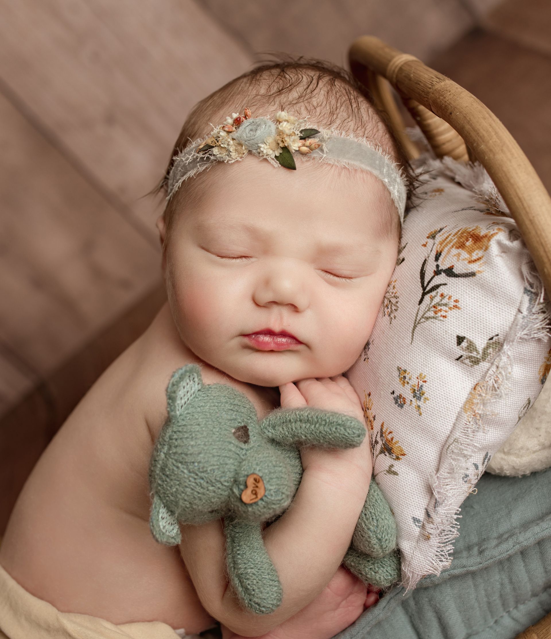 Newborn sleeping in a basket, wearing a floral headband and holding a green stuffed bunny.