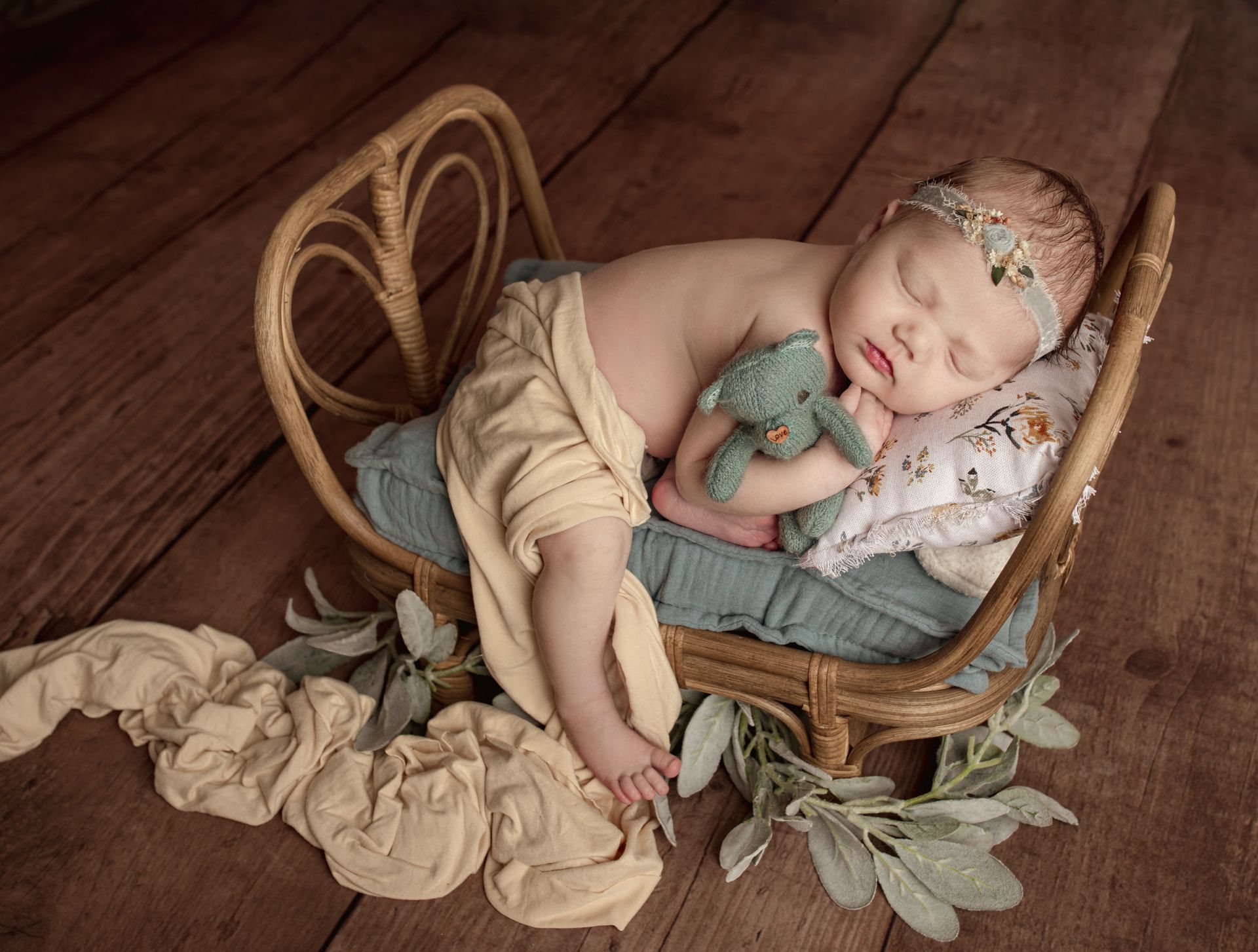 Newborn sleeps in wicker bed with toy. Beige blanket, headband, and wooden floor.