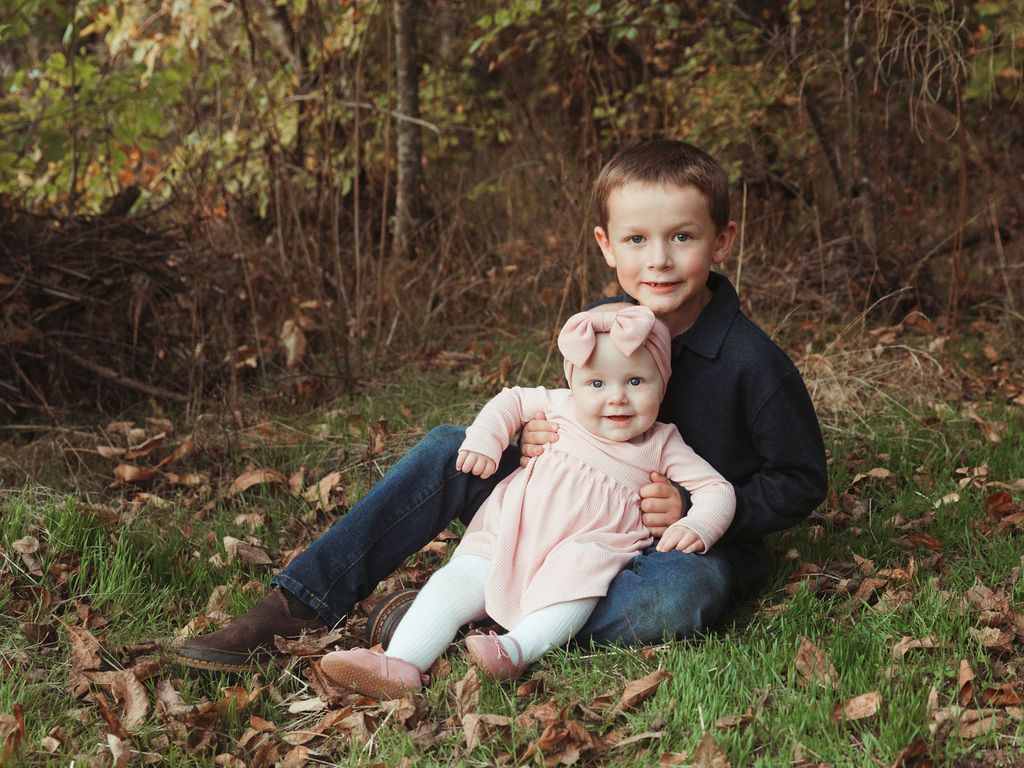 Boy holding a baby, sitting in grass in front of trees. Boy in dark blue shirt, jeans, girl in pink dress, bow.