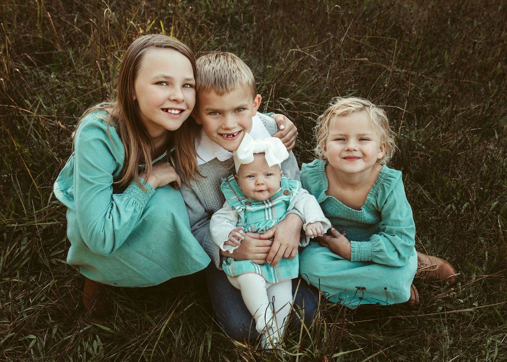 Four children pose in tall grass; two wear teal dresses, one a sweater, and the youngest is held.